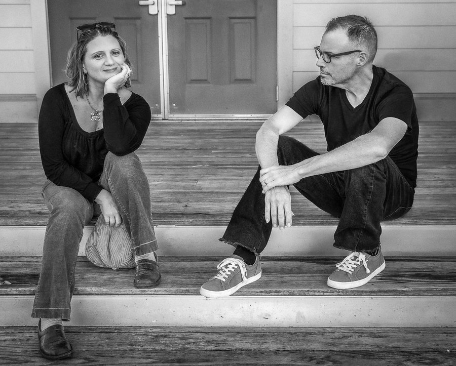 A woman and a man sitting on wooden steps, engaged in a conversation, in front of a door with wooden walls.