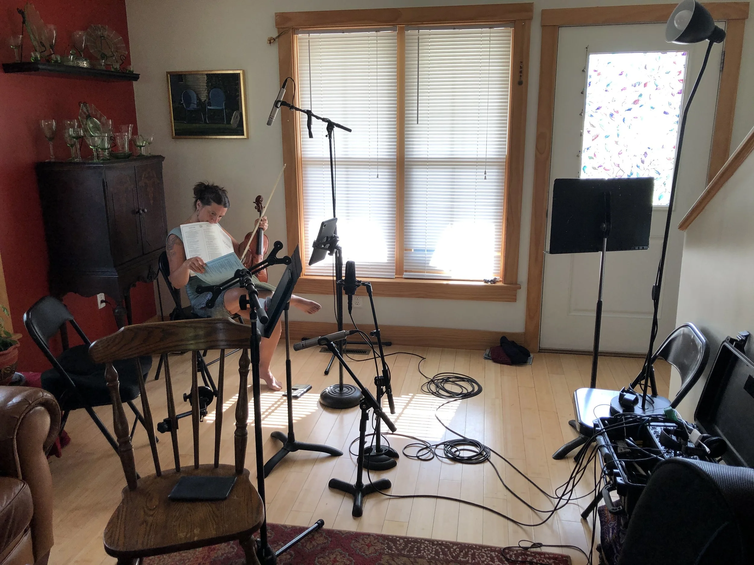 A woman holding a violin going through some sheet music, in a room with some recording equipment getting ready to begin the session.