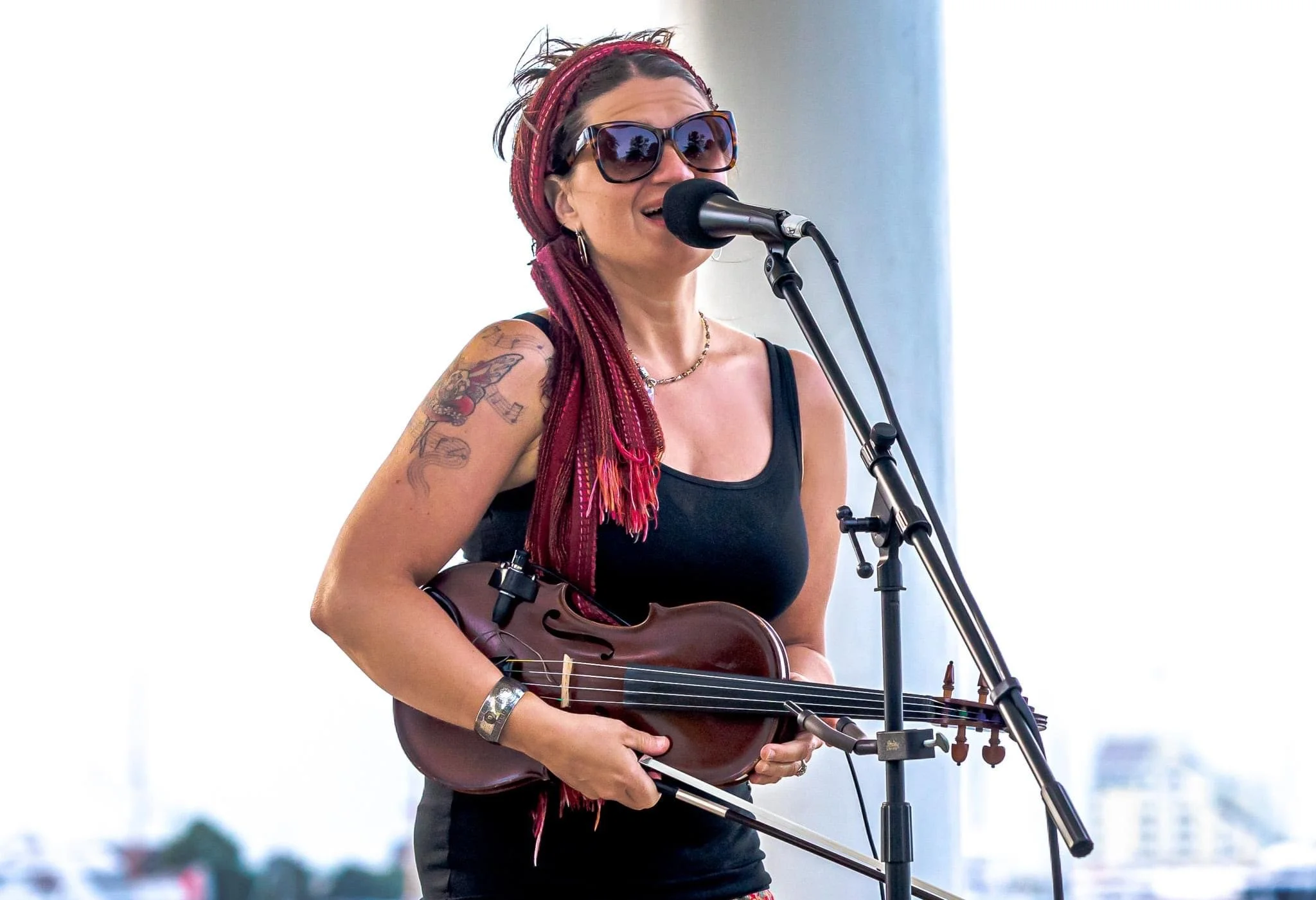 A woman with red dreadlocks, wearing sunglasses, a black top, and a bracelet, holds a brown electric violin and sings into a microphone during a live outdoor performance.