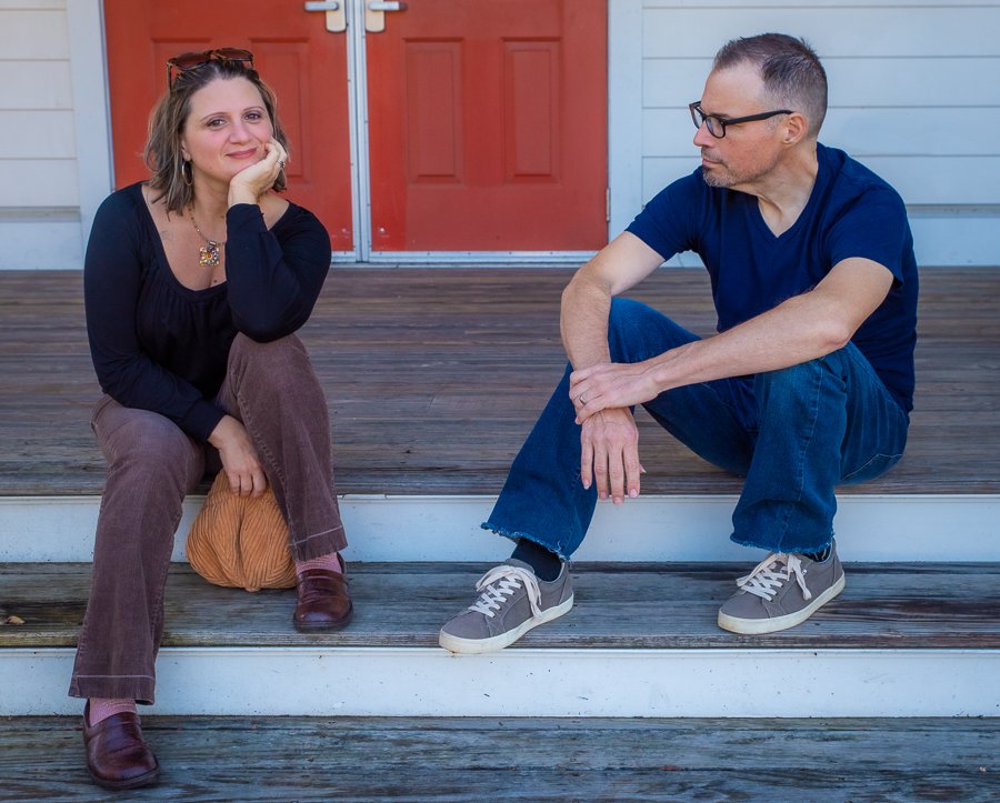 A woman and a man sitting on wooden steps in front of a house with a red door. The woman is smiling, resting her head on her hand, wearing a black top, brown pants, and brown shoes. The man is looking at her, wearing glasses, a navy blue T-shirt, blu