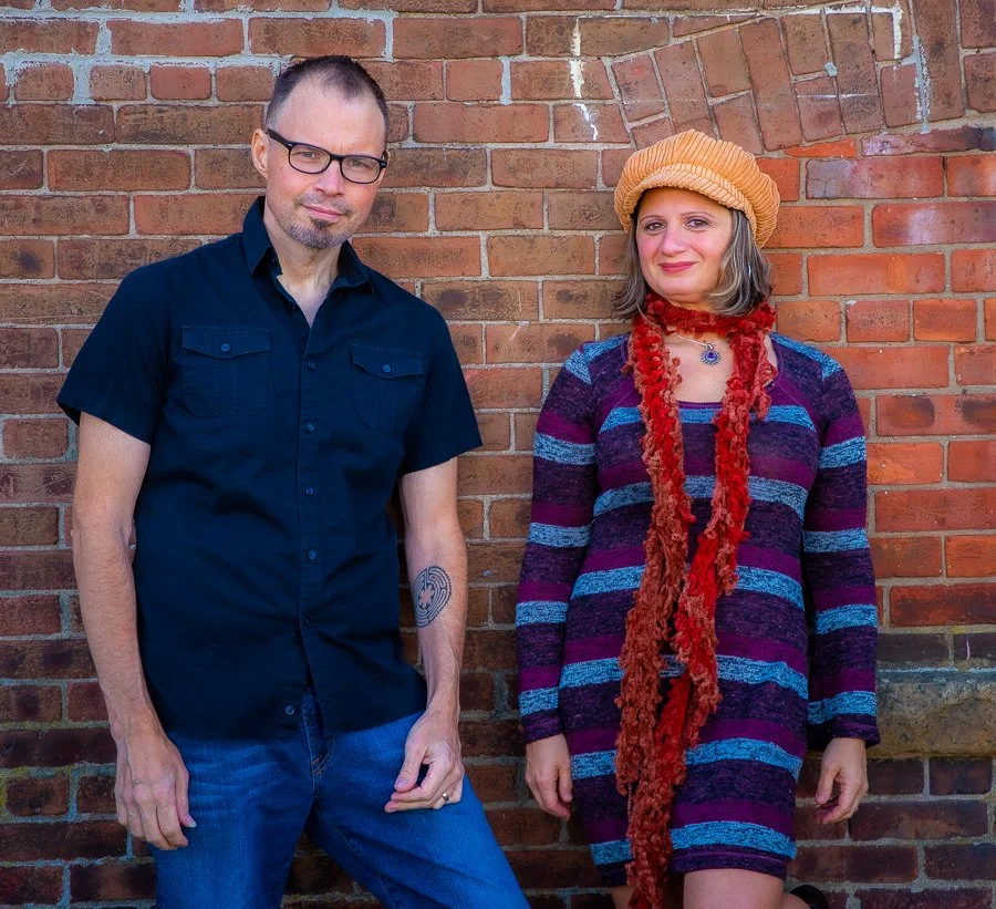 A man and a woman standing next to a brick wall. The man has short dark hair, glasses, a goatee, a black short-sleeved shirt, and jeans. The woman has shoulder-length gray hair, a tan beret, a colorful striped dress, and a red scarf. She is smiling.