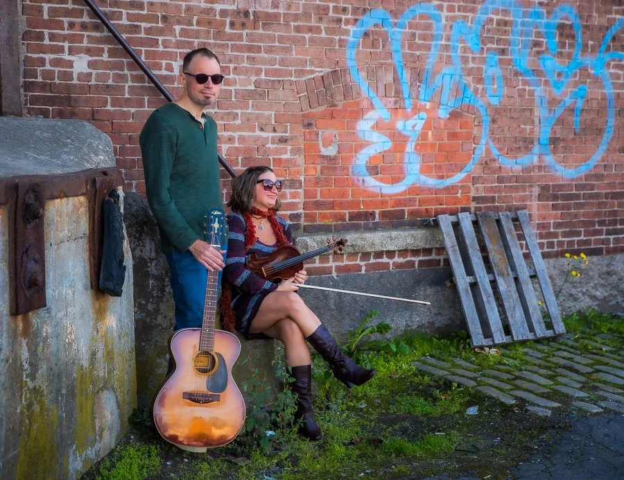 A man and a woman with guitars sitting and standing against a graffiti-covered brick wall in an urban alleyway, with a wooden pallet on the ground beside them.