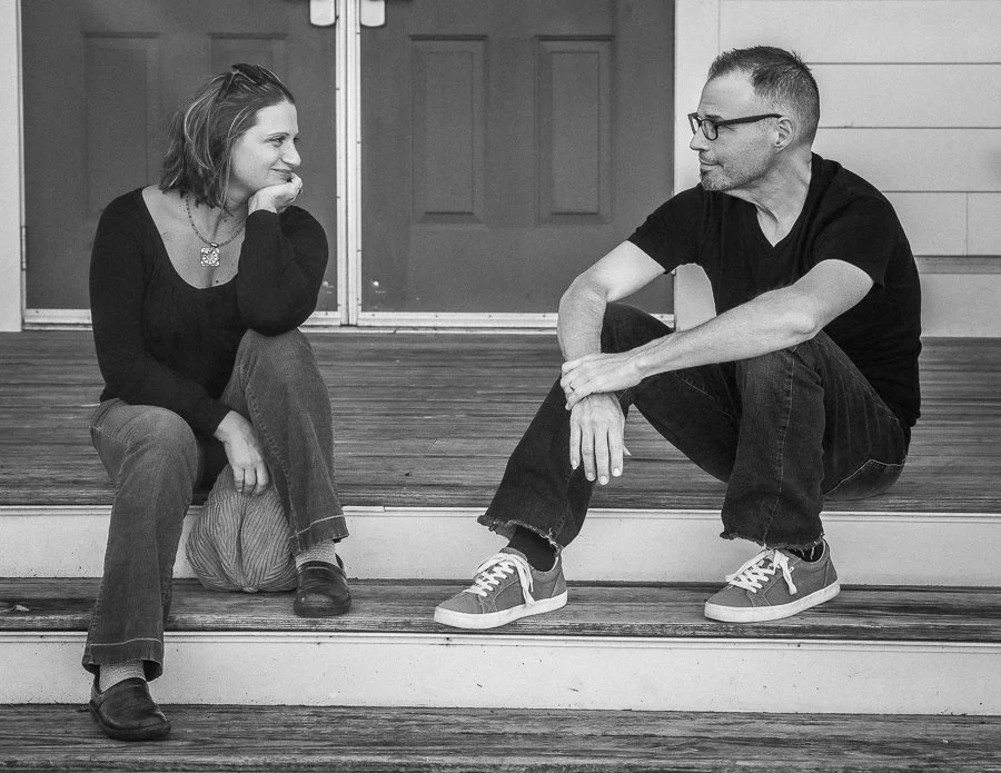 A woman and a man sitting on a staircase facing each other, engaged in conversation, indoors with wooden flooring and closed doors in the background.