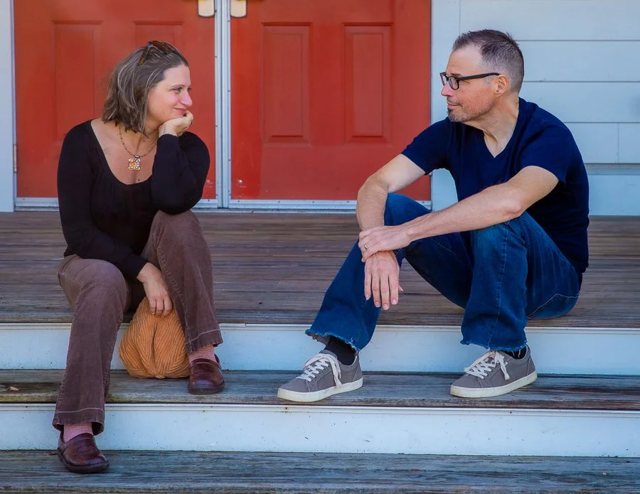 A woman and a man sitting on steps outside, engaged in conversation and smiling at each other.
