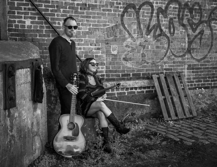 A man with a guitar and a woman with a violin sitting  and standing against a brick wall with graffiti, wearing sunglasses, in black and white.