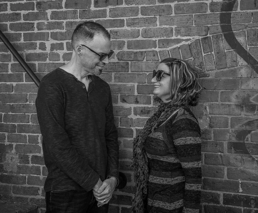 A man and woman stand facing each other, smiling, against a brick wall. Both are wearing glasses and casual clothing.