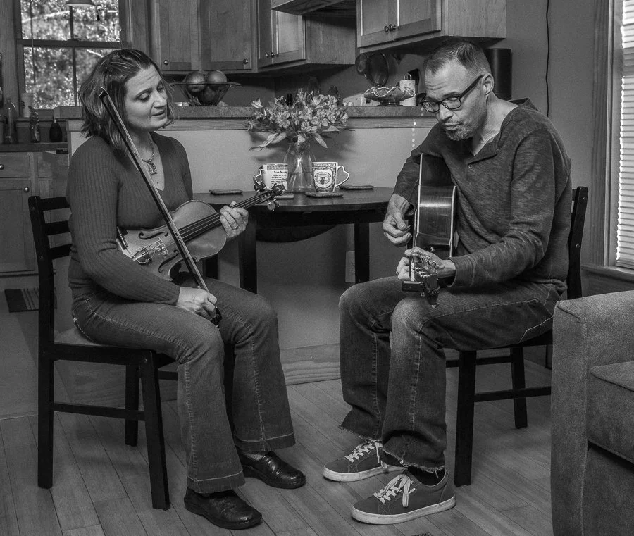 A woman and man playing guitars together in a cozy kitchen, seated on chairs facing each other, a table with a flower arrangement in the background.