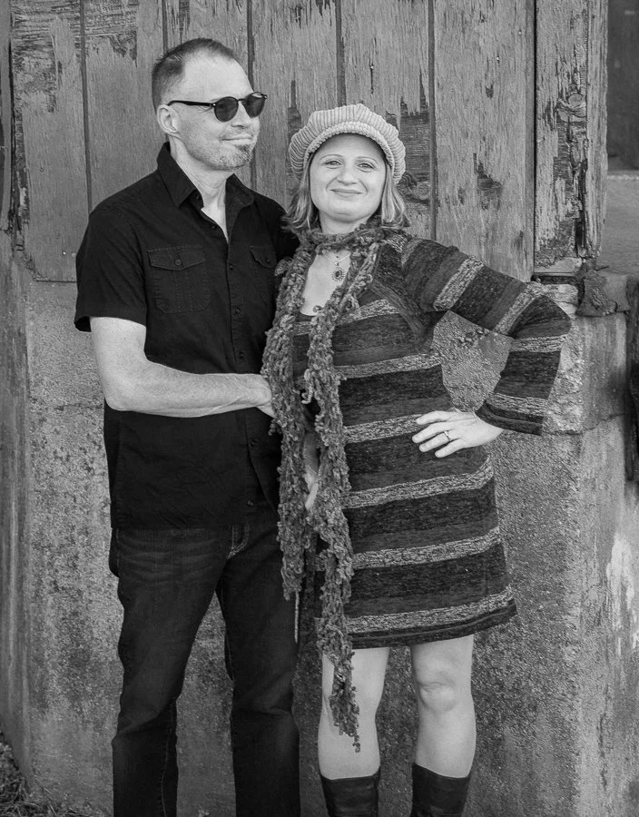 A man and a woman standing together outdoors in front of a wooden wall, smiling at the camera. The man is wearing sunglasses, a short-sleeved button-up shirt, and jeans. The woman is wearing a patterned dress, a knit hat, a scarf, and boots.