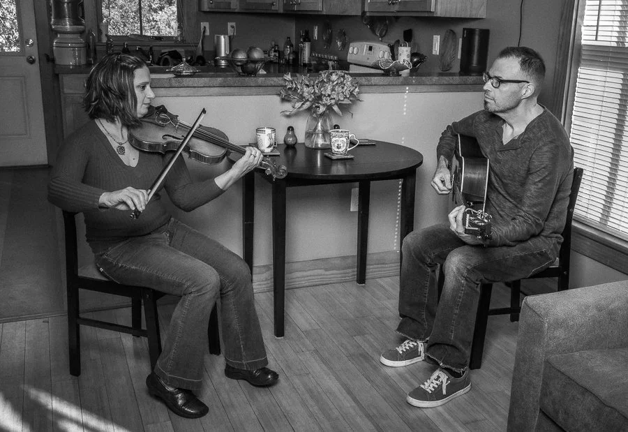 A woman playing a violin and a man playing an acoustic guitar in a home kitchen and dining area.