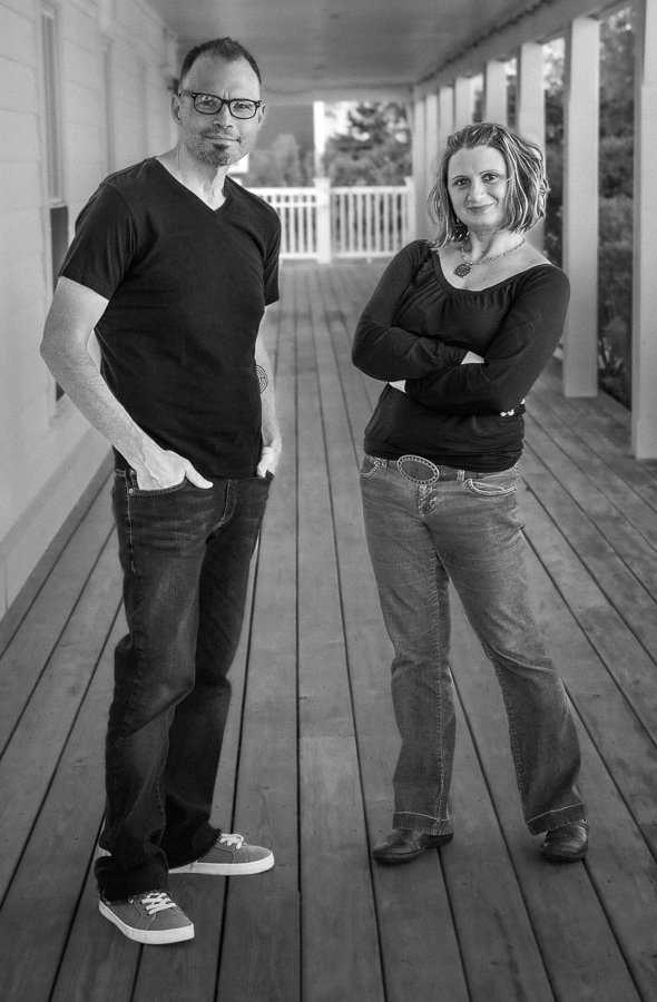 A man and woman stand on a porch, facing the camera, with the man wearing glasses and a black t-shirt, and the woman crossing her arms in a black top and jeans.