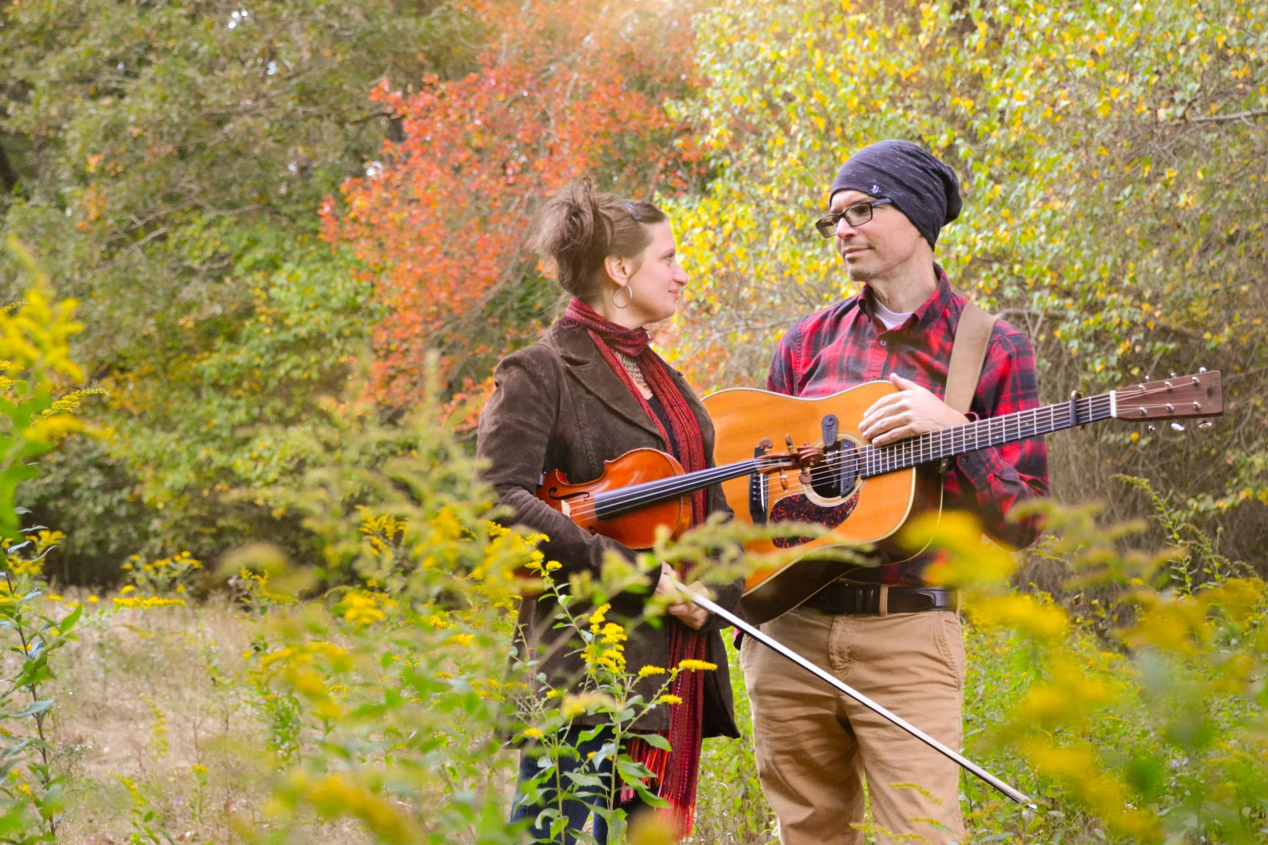 A man and woman holding their guitar and violin outdoors in a colorful autumn forest, looking at each other.