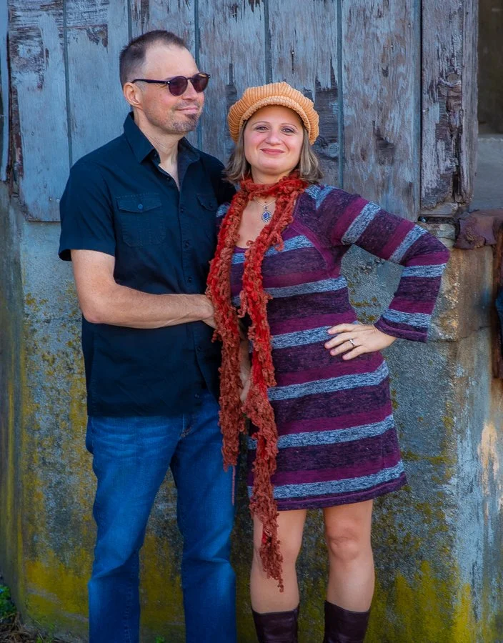 A man and a woman standing and smiling in front of a rustic, weathered blue and gray wooden wall.