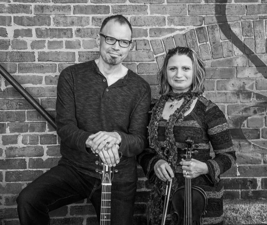 A man and woman standing in front of a brick wall, each holding a musical instrument. The man is wearing glasses and a dark long-sleeve shirt, holding a guitar. The woman has shoulder-length hair, is wearing a patterned sweater with a scarf, and is h