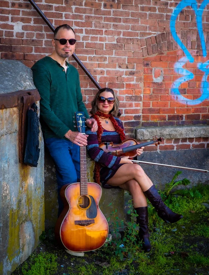 A man and woman with musical instruments leaning against a brick wall with graffiti. The man is holding an acoustic guitar, and the woman is sitting with a violin and bow. Both are wearing sunglasses, and the woman has a red scarf around her neck.