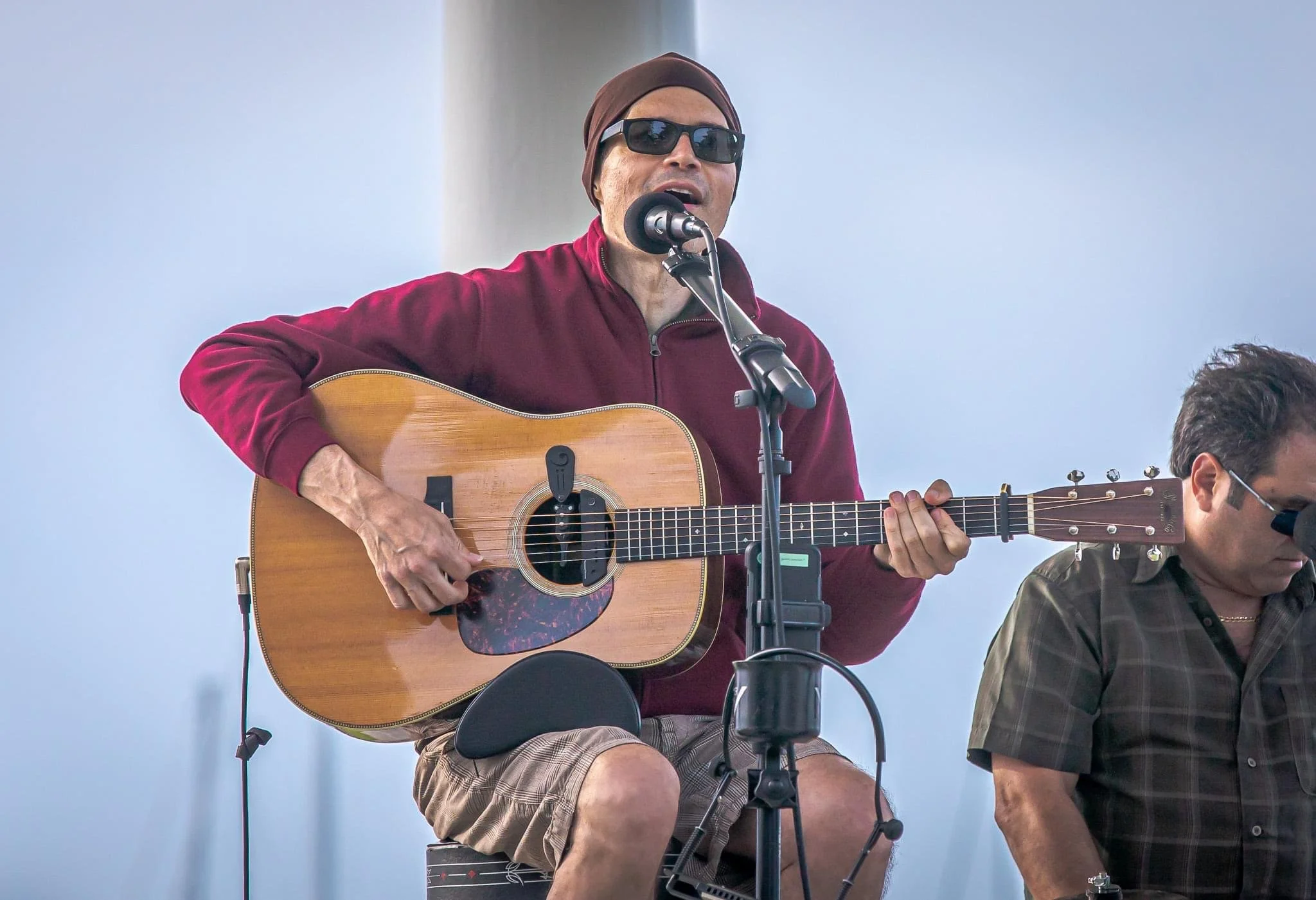 Man wearing sunglasses and a brown headscarf playing an acoustic guitar and singing into a microphone.