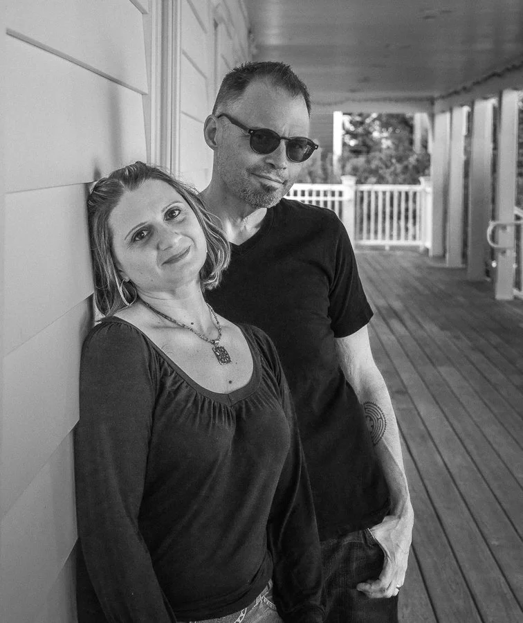 Black and white photo of a woman and man leaning against a wall on a porch. The woman has short hair, hoop earrings, and a necklace, wearing a dark top. The man has short hair, sunglasses, a beard, a tattoo on his arm, and is wearing a black T-shirt.