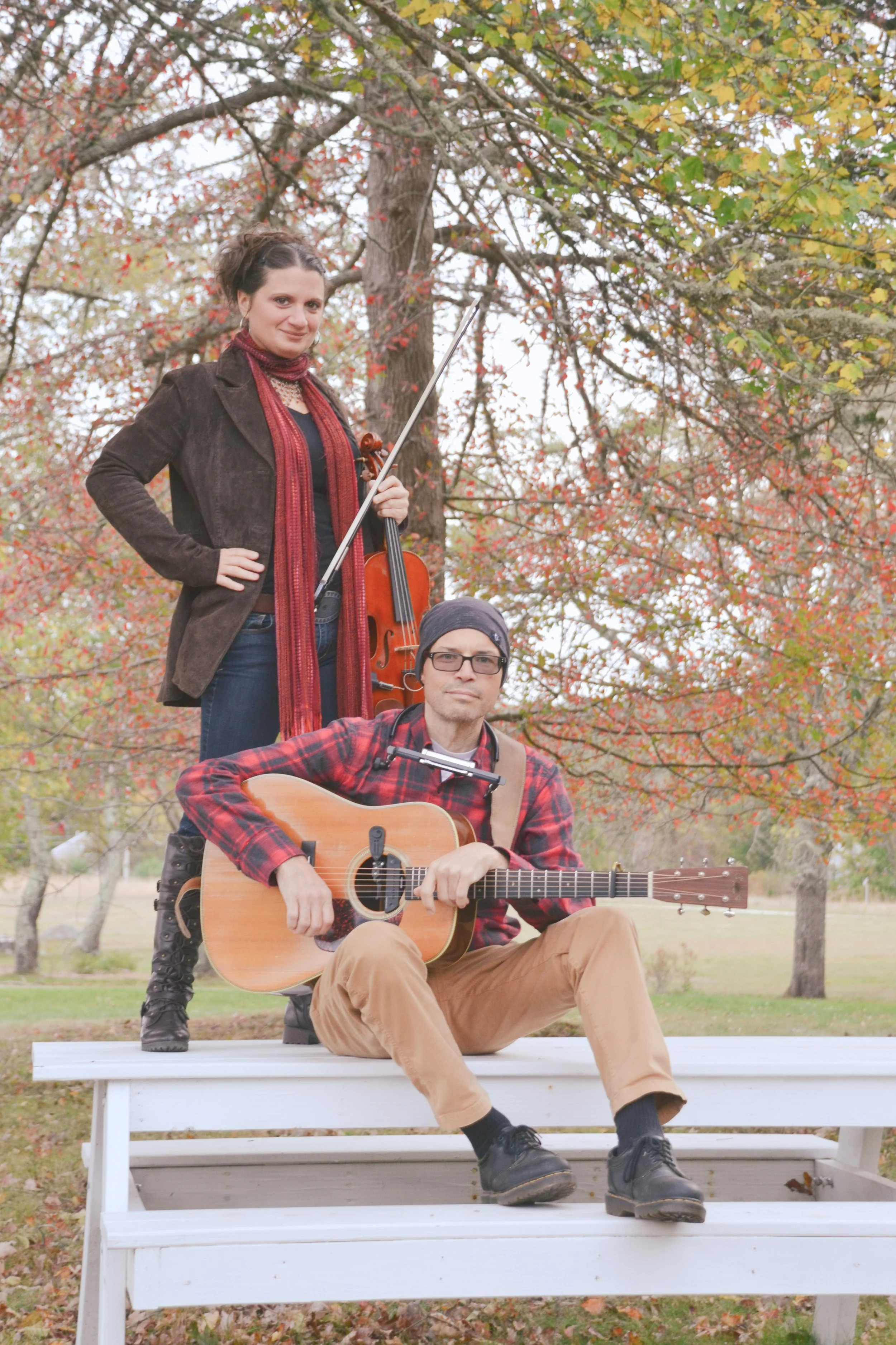 A man playing guitar and a woman holding a violin outdoors in fall, with trees displaying colorful autumn leaves.