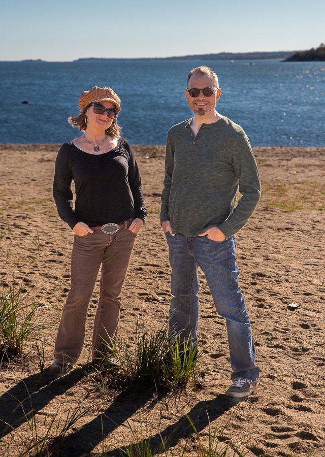 A man and a woman standing on a sandy beach near the water, both wearing sunglasses and smiling, with the woman in a black top, brown pants, and a straw hat, and the man in a green shirt and jeans, with a calm ocean and a distant shoreline in the bac