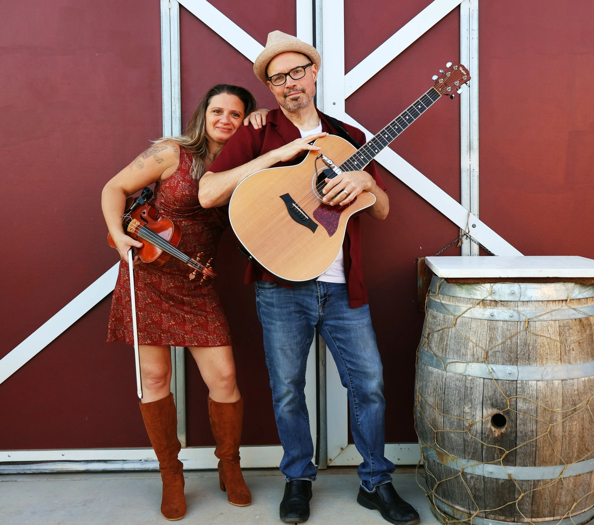 Virginia James Duo standing in front of a red barn door with white trim holding a guitar and a violin.