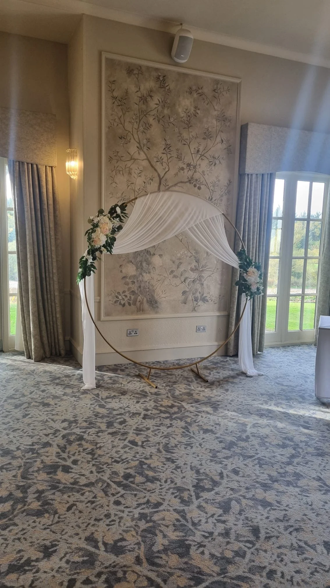 A floral wedding arch with white drapes and pink and white flowers, set up indoors near large windows with curtains, on a patterned carpet.