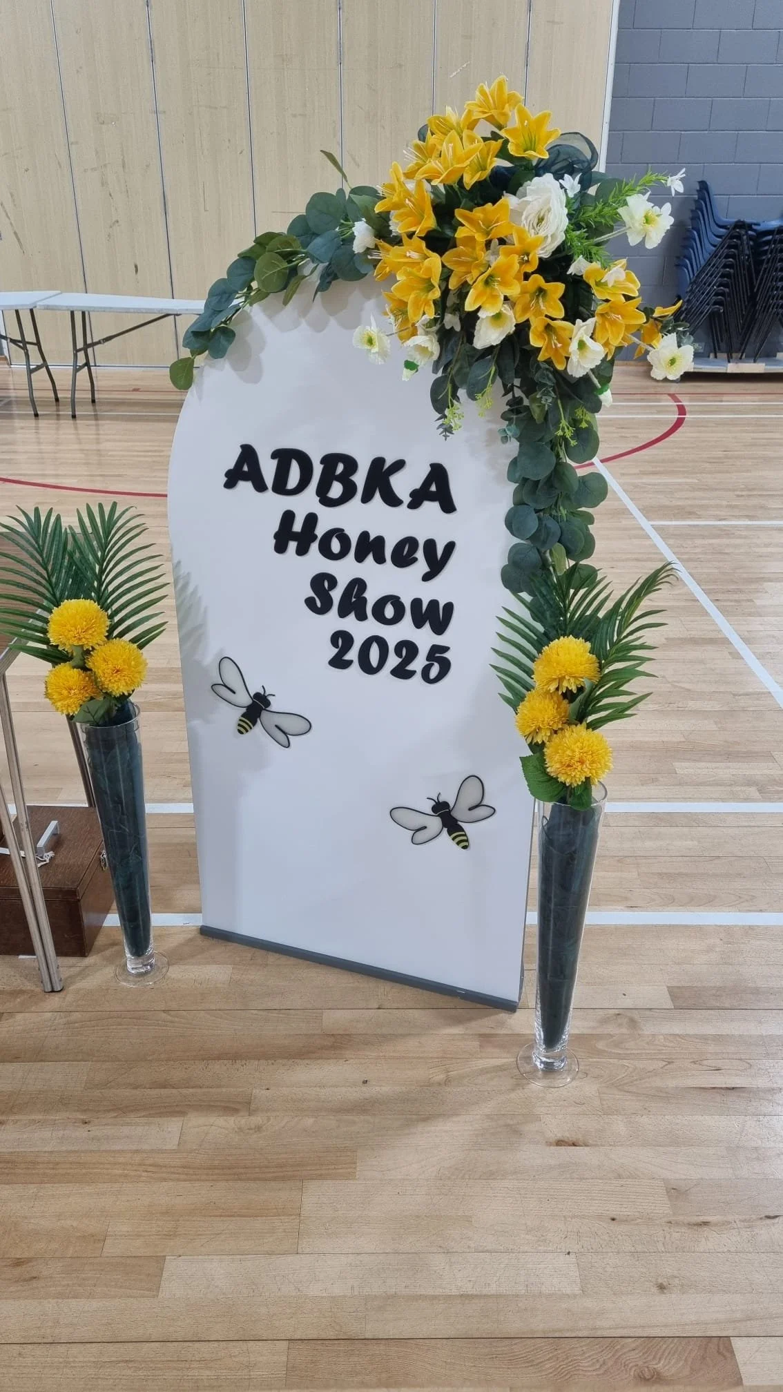 A white sign with black text that reads "ADBA Honey Show 2025" decorated with yellow and white flowers and green leaves, flanked by tall glass vases with yellow flowers and green foliage, set on a wooden floor in a gymnasium.