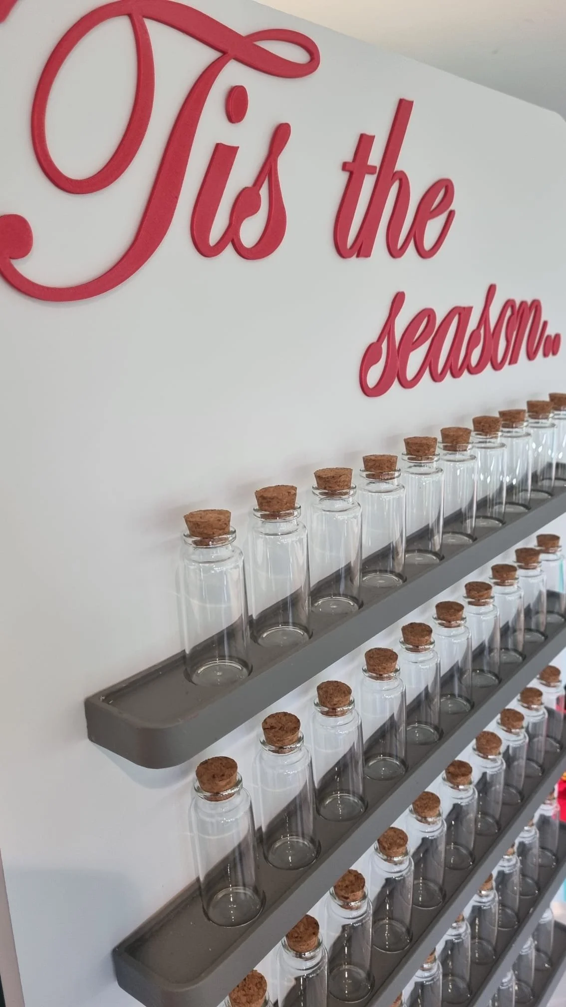 A wall decorated with a red cursive sign that says 'Tis the season...'. Beneath the sign, there are multiple glass bottles with cork stoppers arranged on gray shelves.