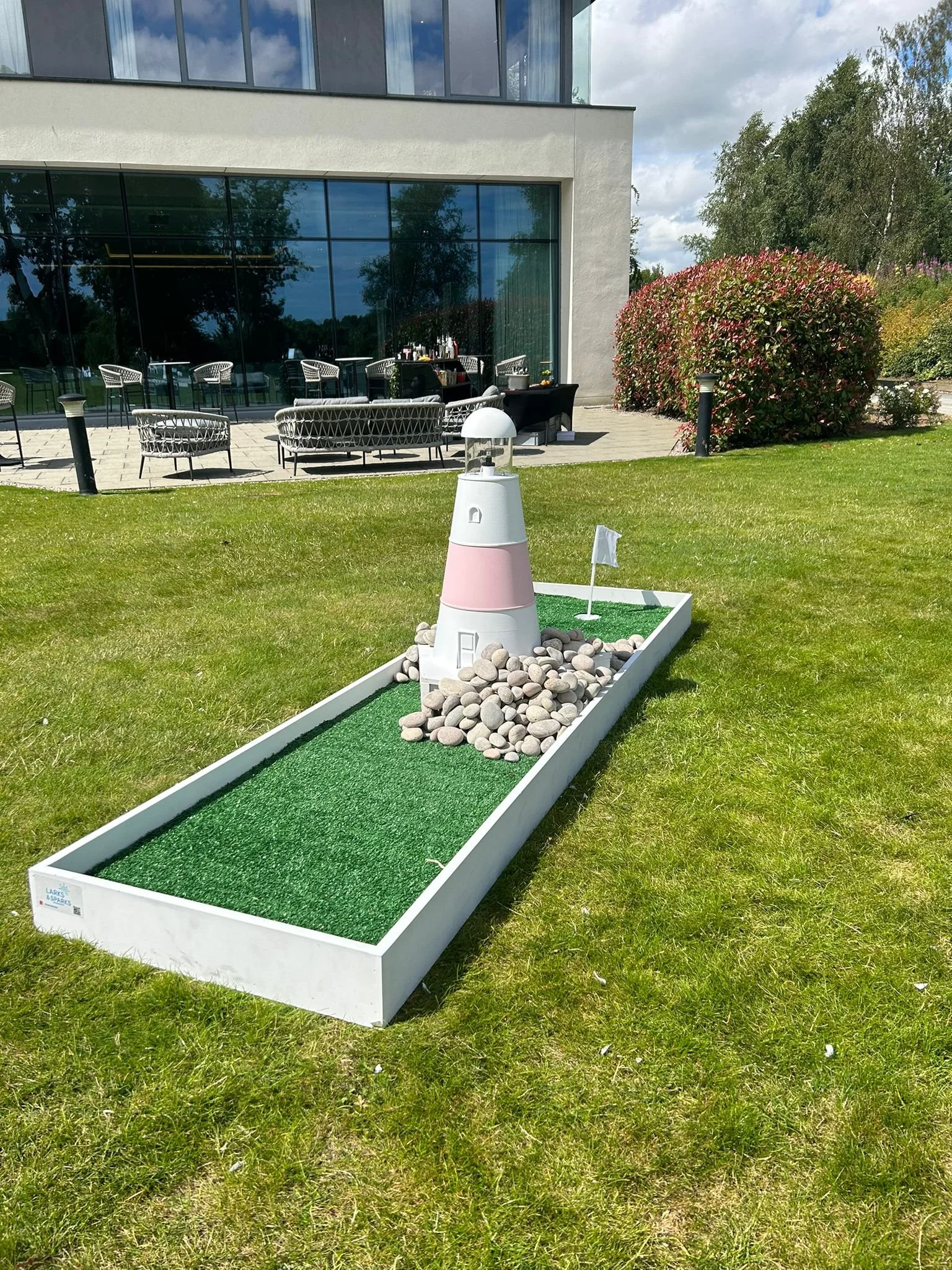Miniature golf hole with a lighthouse-themed obstacle, rocks, and artificial turf, set on a well-maintained lawn outside a modern building with outdoor seating.