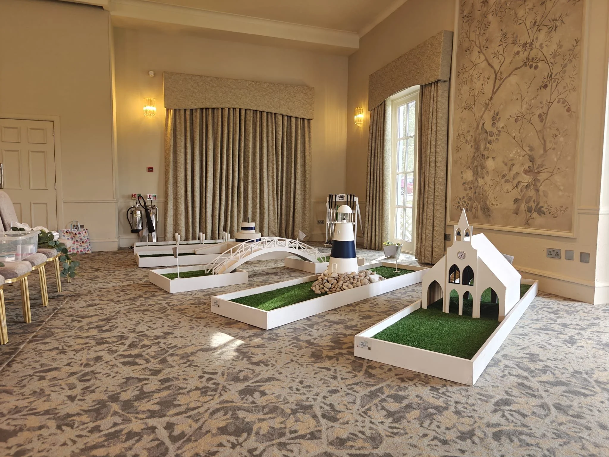 Indoor room with decorative display including tiered miniature landscape, small bridge, lighthouse, and church models with artificial grass, set on white platforms, with ornate curtains, patterned carpet, chairs, and wall sconces.