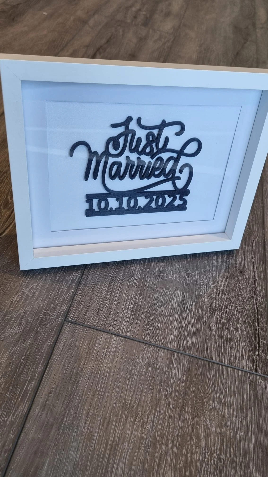 White picture frame with a decorative sign reading 'Just Married' and the date '10.10.2023' on a wood floor.
