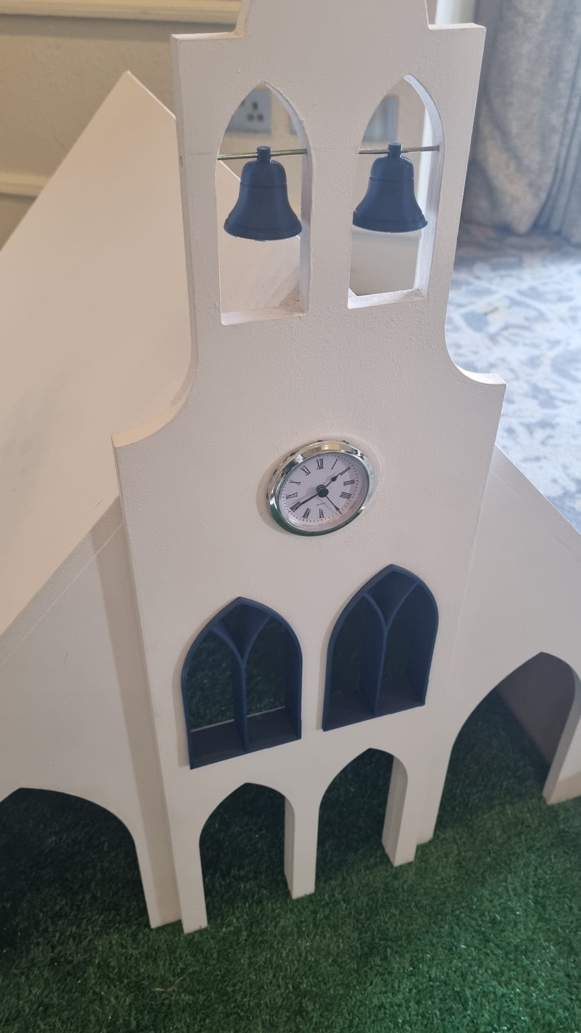 A miniature white model of a church with blue bells, a clock, and blue windows, placed on a green surface.