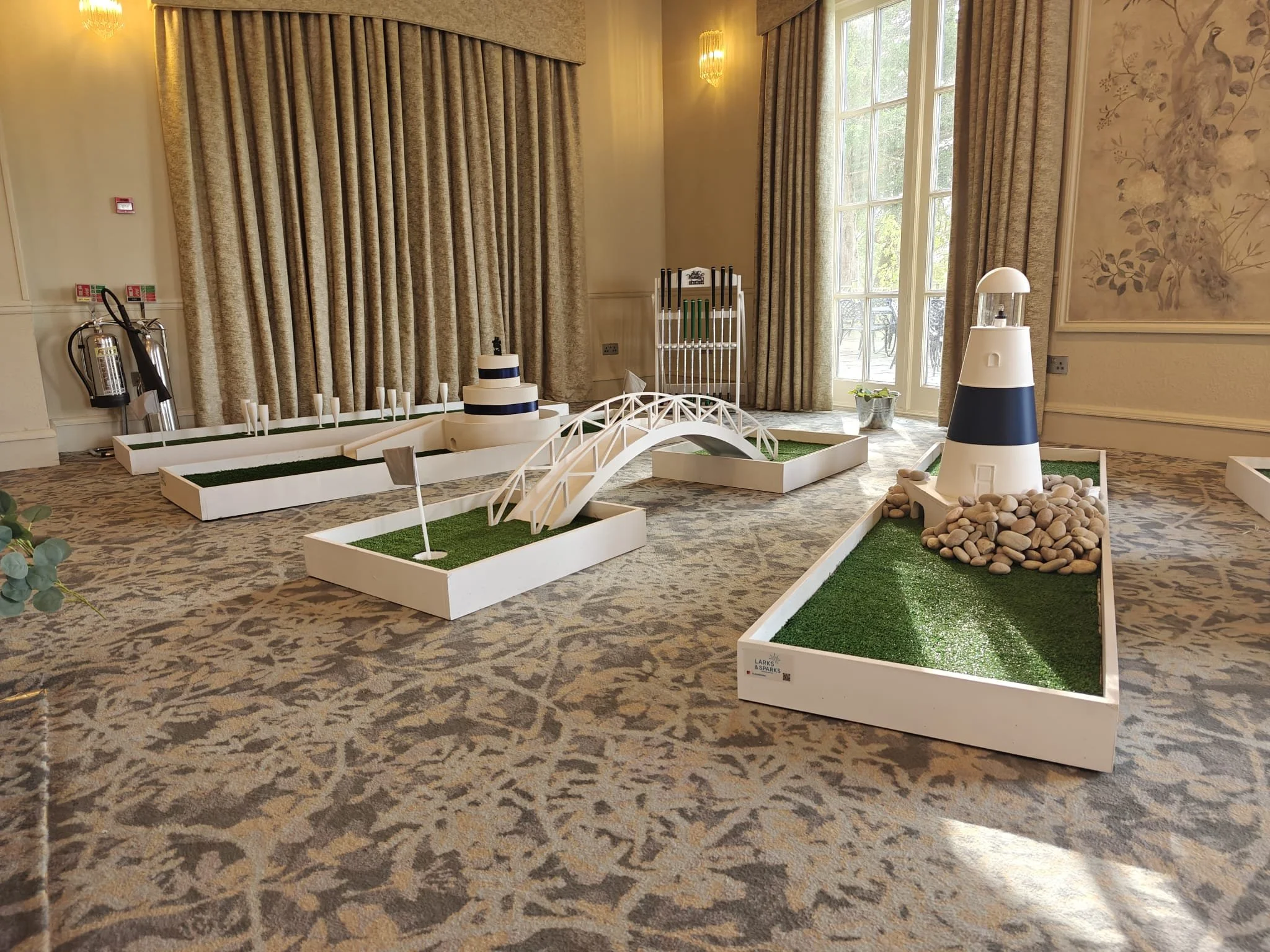 Miniature golf course setup indoors with a lighthouse, bridge, and various holes, on a patterned carpeted floor near large windows and curtains.