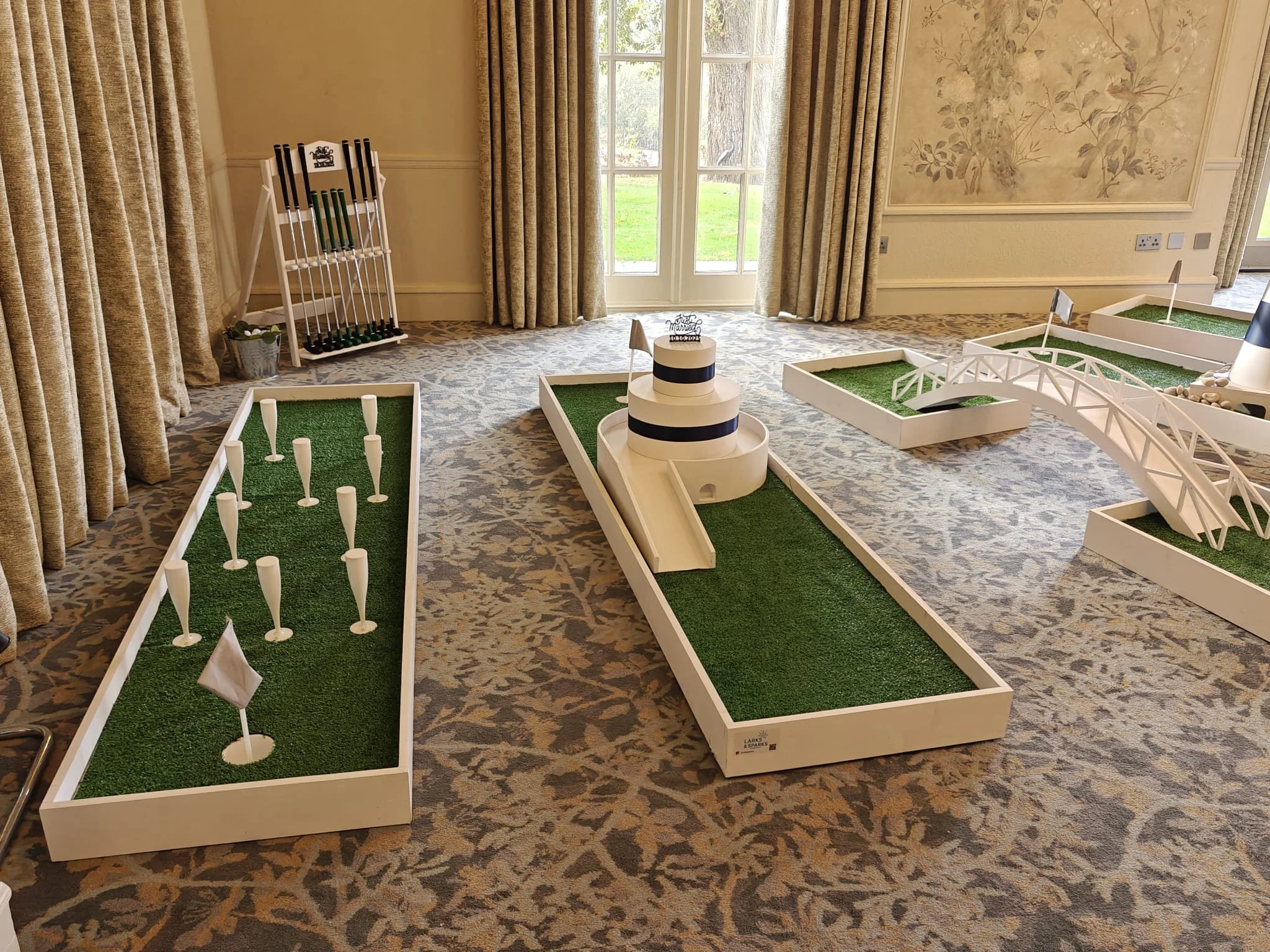 Mini golf setup with artificial turf hazards inside a room, including a small putting green, a bridge, a ramp, and obstacles like golf clubs and flags.