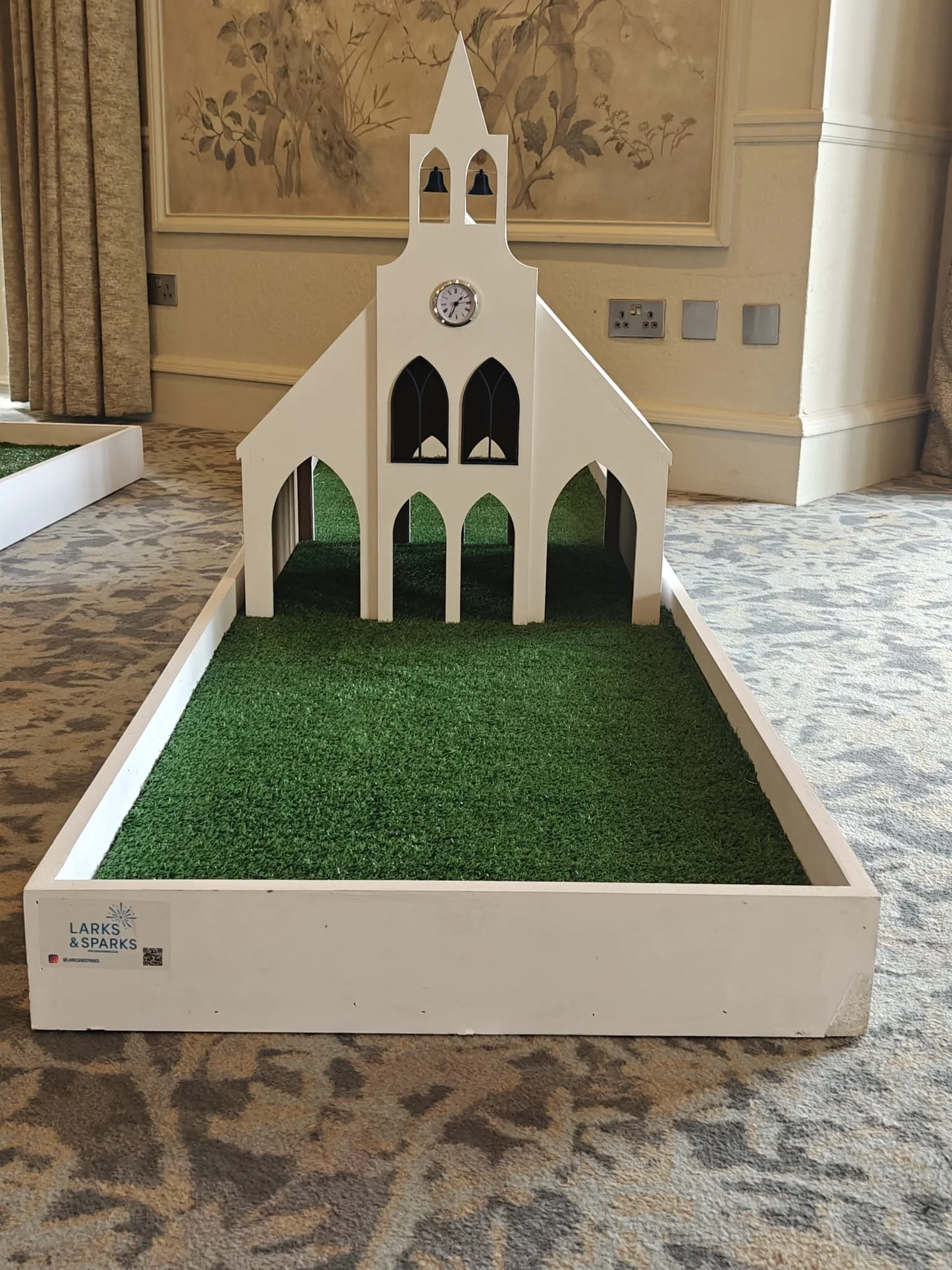 A miniature model of a white church with a clock and black bell towers on a rectangular patch of green artificial grass, placed on a patterned carpeted floor in a room with beige walls and framed wall art.