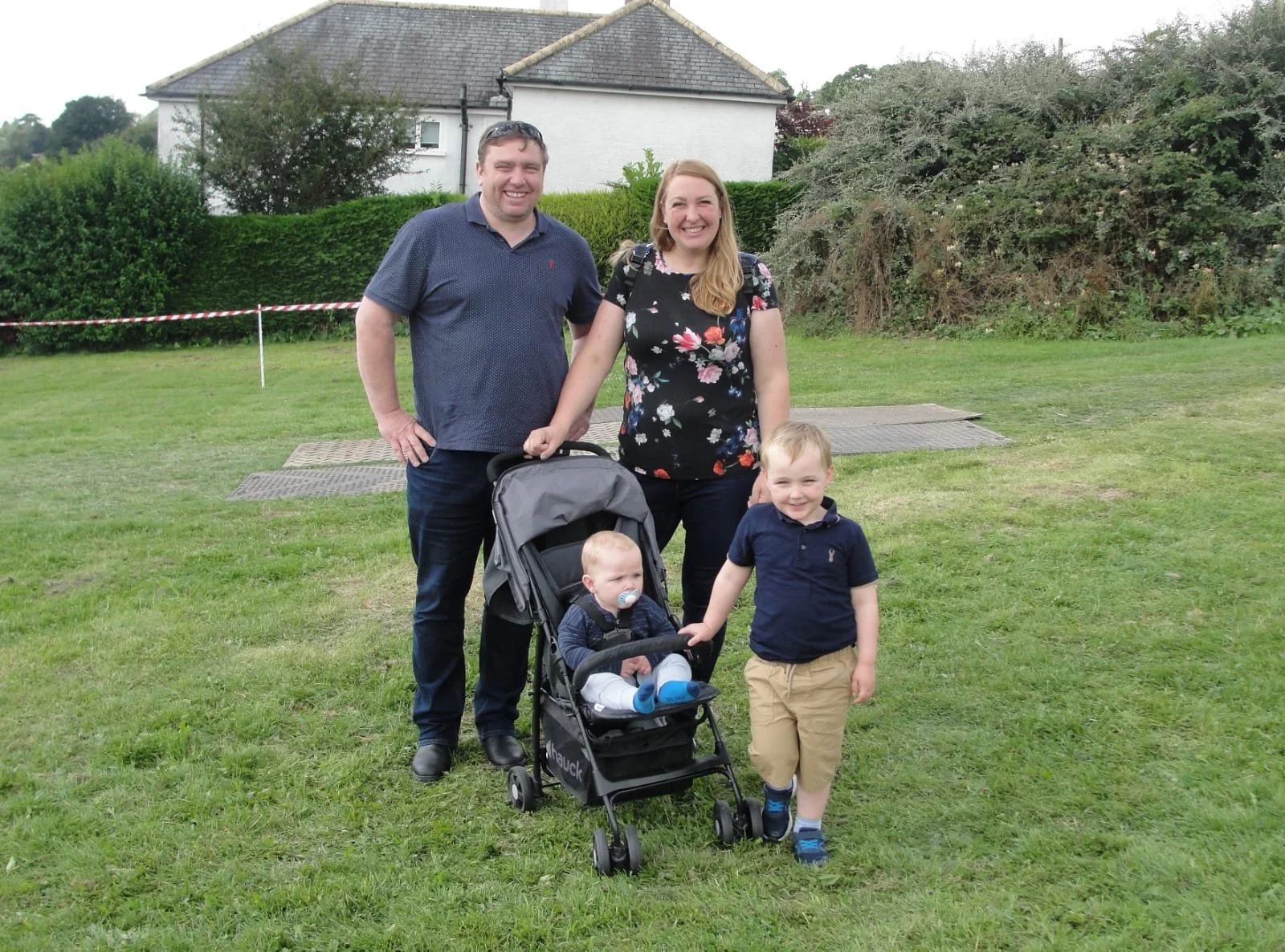 A family of four outdoors on a grassy field, smiling for a photo. The father is standing on the left, wearing a blue polo shirt and jeans. The mother is standing on the right, wearing a black floral top and black pants. The toddler is sitting in a stroller with a pacifier, wearing a dark jacket and white pants. The young boy is standing next to the stroller, wearing a navy blue polo shirt and khaki shorts.