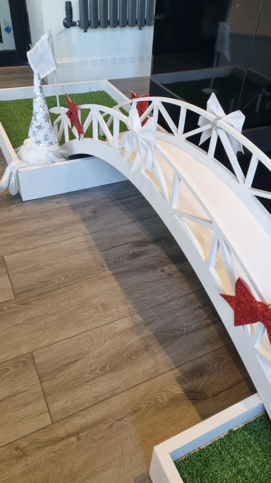 White decorative bridge with holiday bows and a Christmas hat on a small gnome figure, placed over artificial grass on a wooden floor.