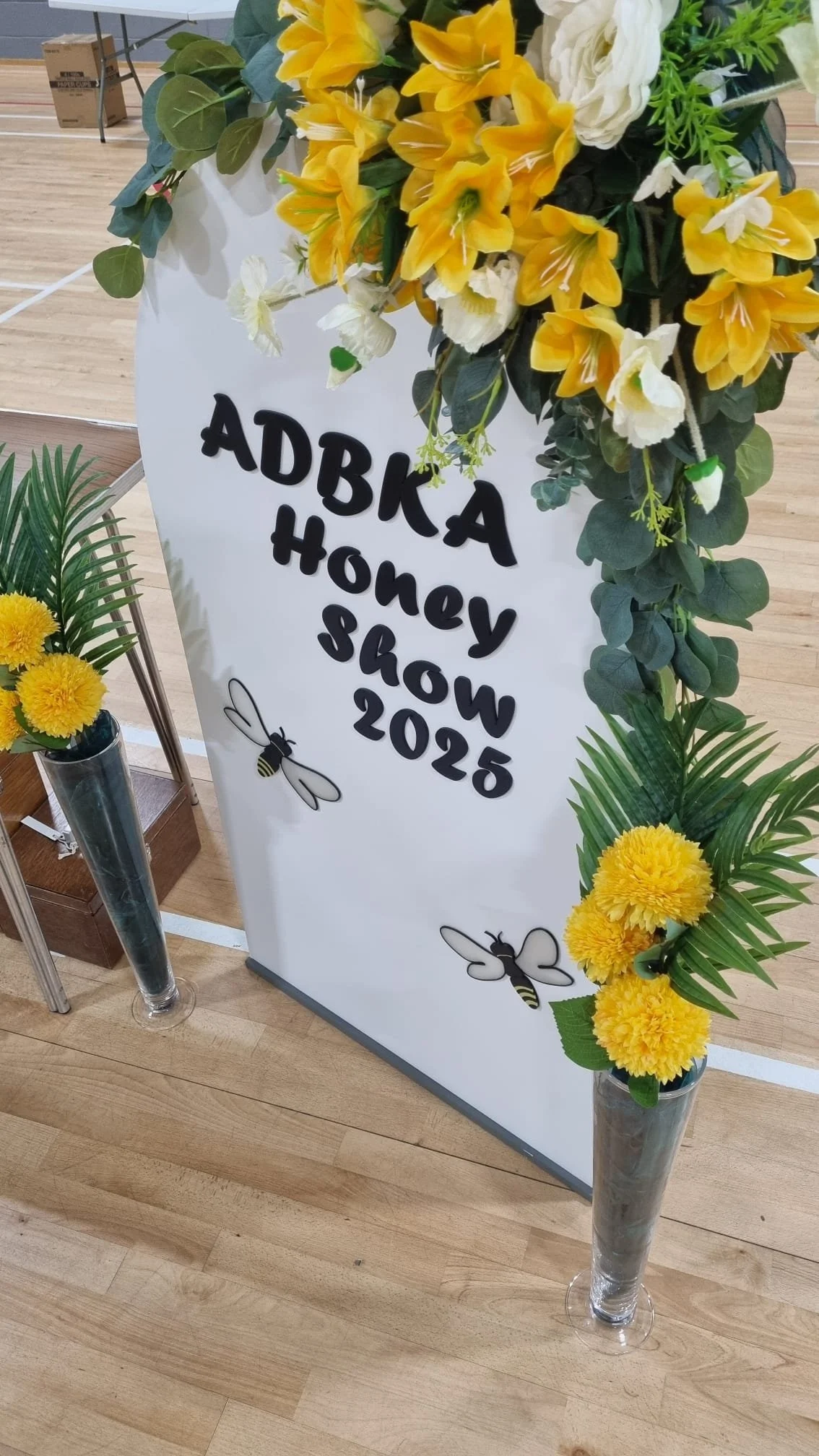 Floral display with yellow and white flowers around a sign that reads 'ADKAA Honey Show 2025' with illustrations of bees.