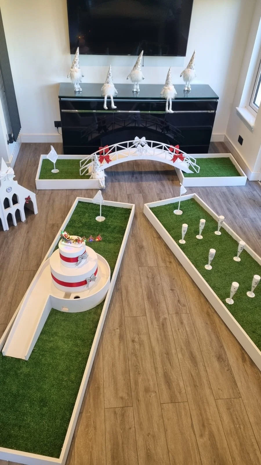 Miniature golf course with holiday decorations including a small white bridge with red bows, a three-tiered white cake with red ribbons and bows, and figurines of gnomes in festive hats on a black cabinet, all set in a living room with wooden floorin