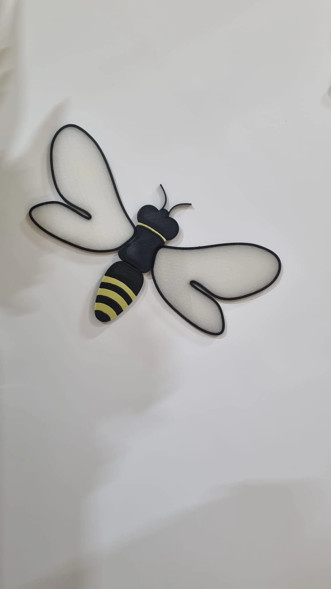 A fabric bee decoration with black and yellow striped abdomen, black head and thorax, and white wings outlined with black, lying on a white surface.