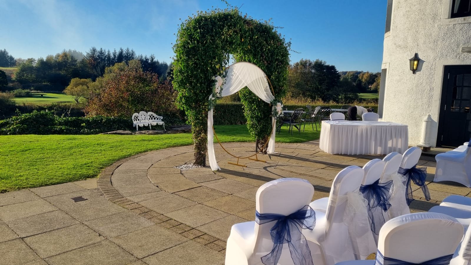Outdoor wedding ceremony setup with white chairs decorated with navy blue ribbons, a white draped arch with greenery, and a scenic backyard landscape with trees and a bench under a clear blue sky.