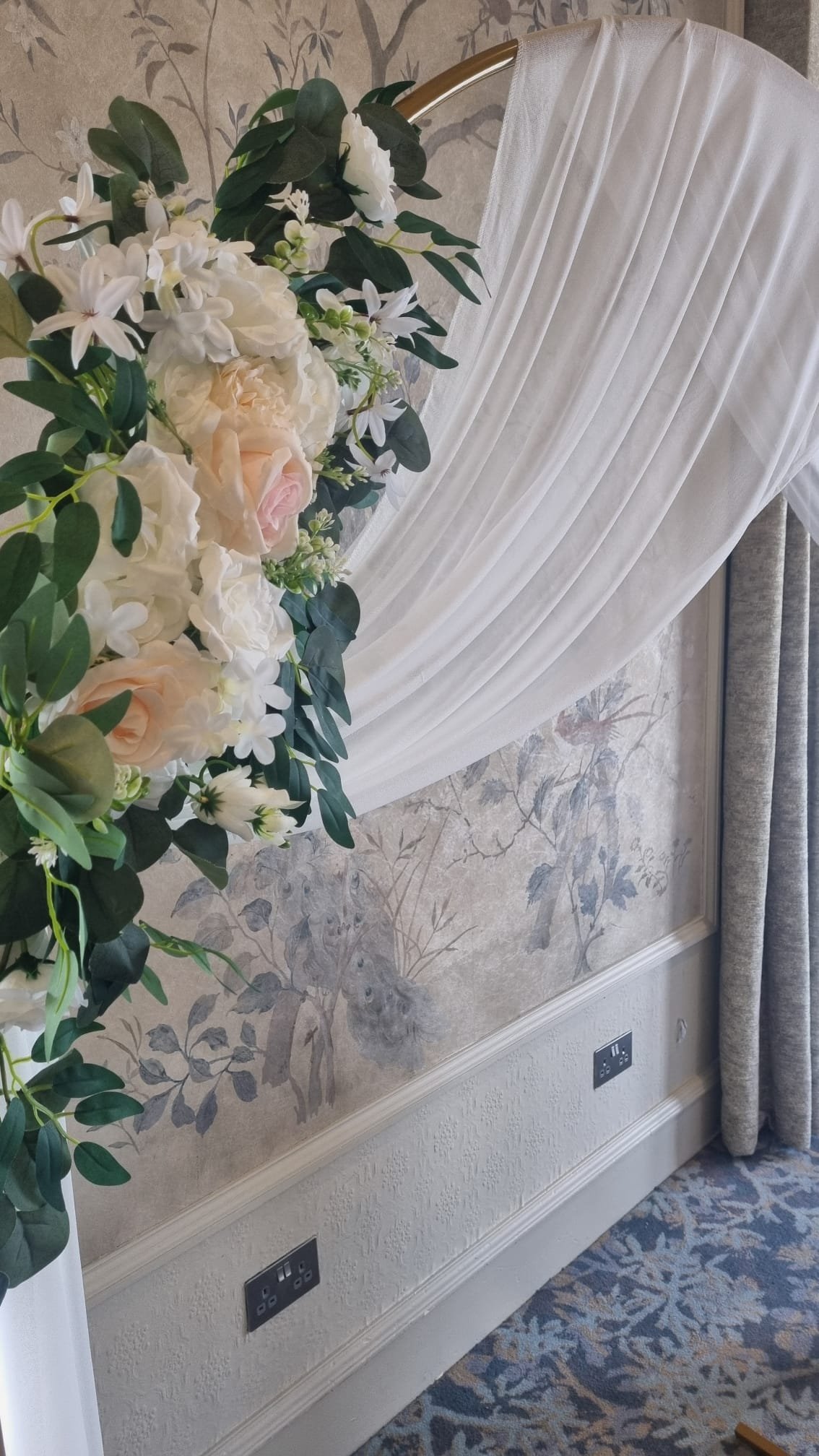 Decorative wedding arch with white drape and floral arrangements of white and blush roses, white lilacs, and green foliage, set against a nature-themed wall with prairie and fox print.