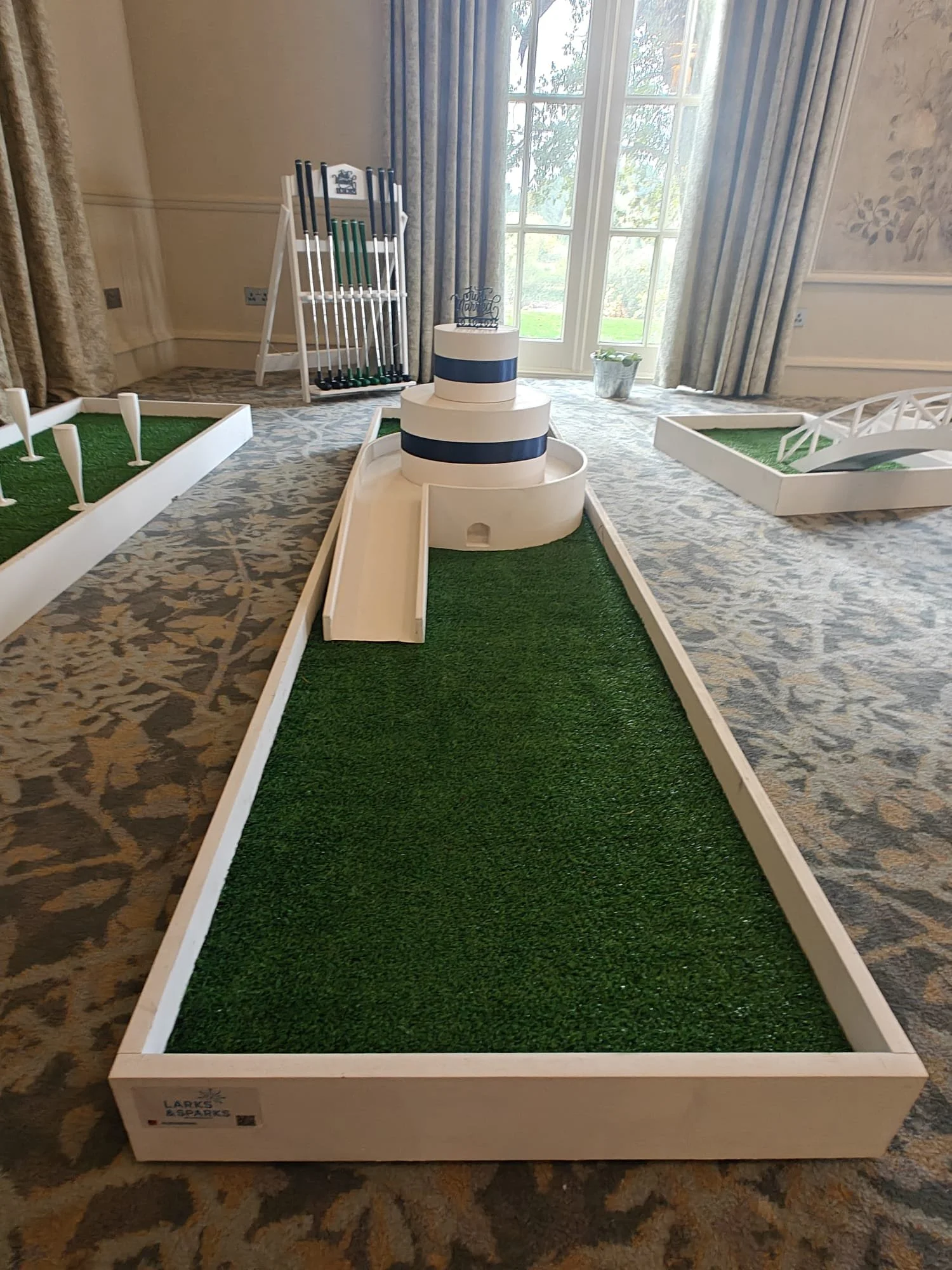 Miniature golf holes with artificial grass and ramps, set up indoors near large windows with curtains.