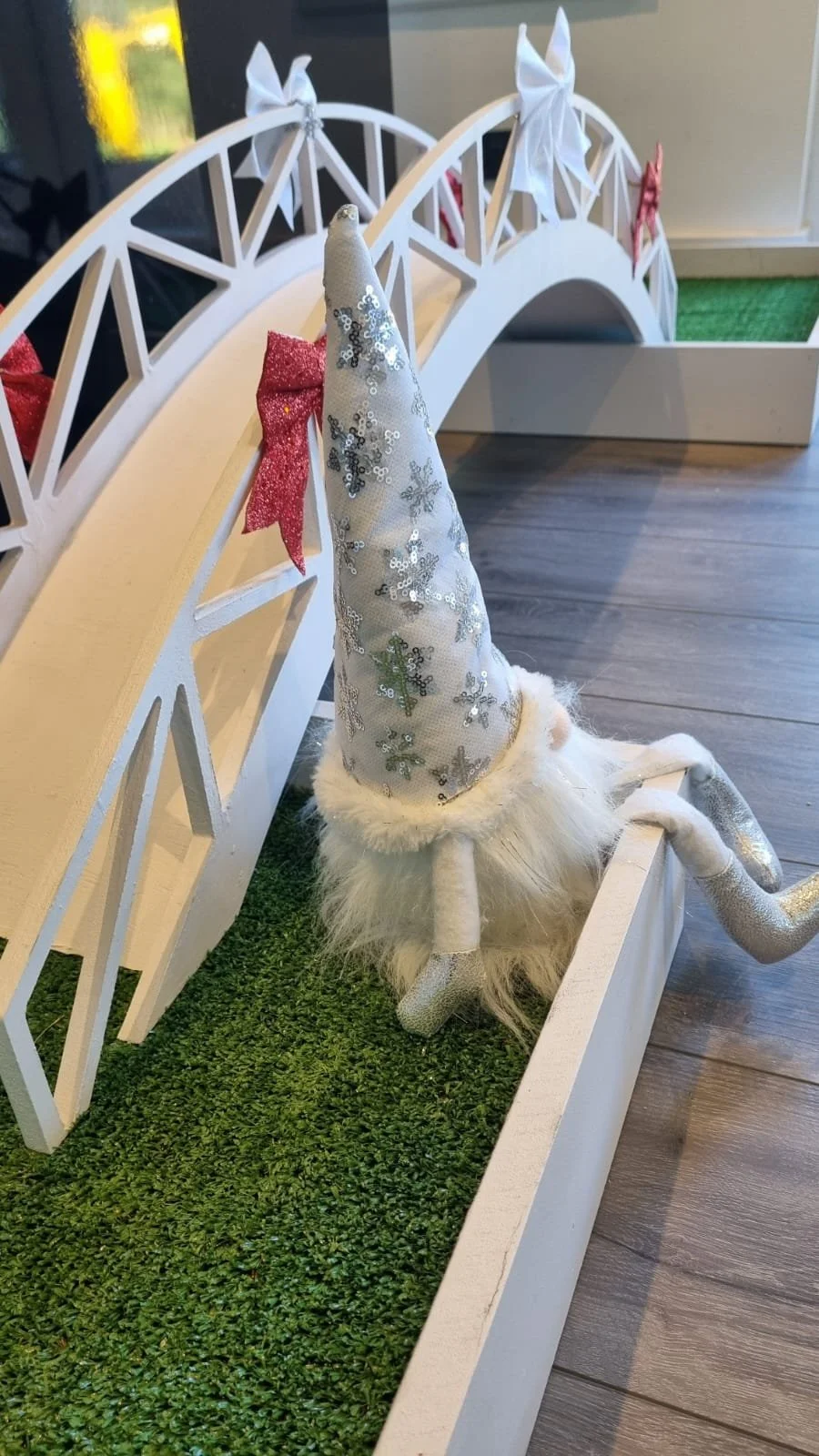 Decorative Santa hat with sequins and a plush beard, leaning against a white picket fence with bows, on a patch of green turf.