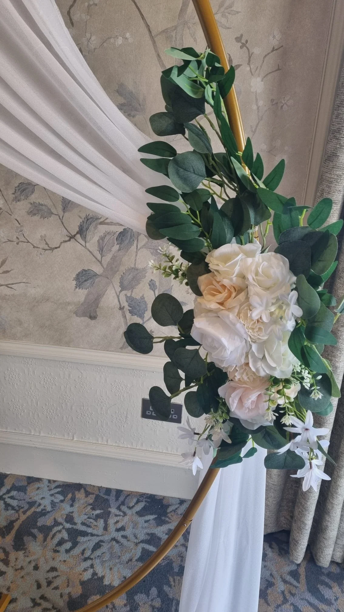 Flower arrangement with white roses, lilies, and green leaves on a gold stand.