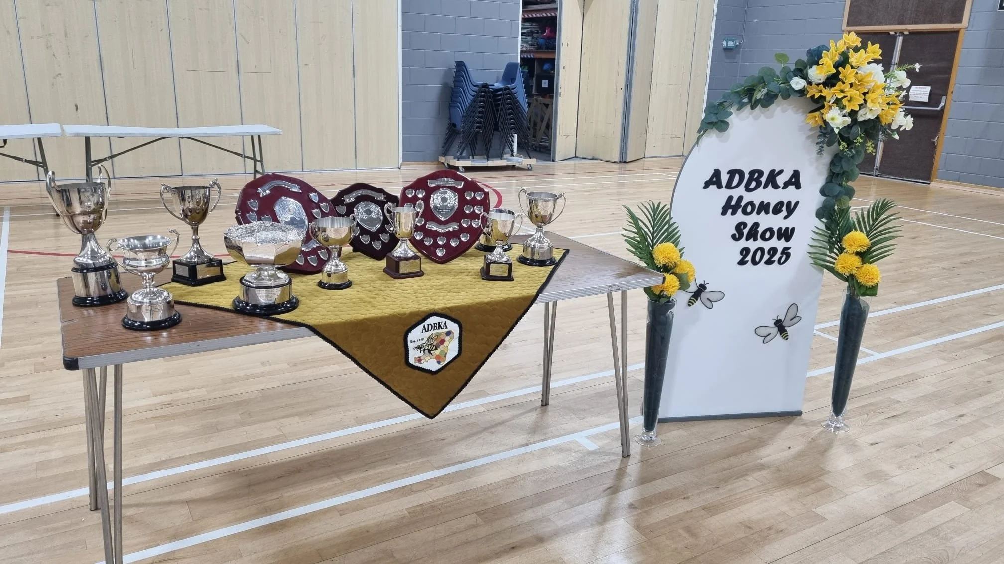 Awards display table with trophies and plaques at the ADKBA Honey Show 2025, decorated with a floral arrangement and a sign in a gymnasium.