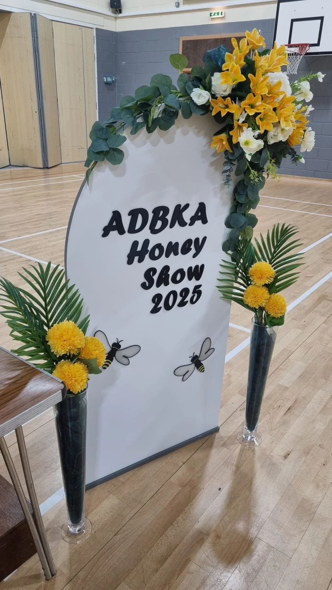 A white, arc-shaped signboard decorated with yellow and white flowers, green leaves, and bee illustrations reads 'ADBA Honey Show 2025,' placed in a gymnasium with wooden floors and basketball hoop in the background.