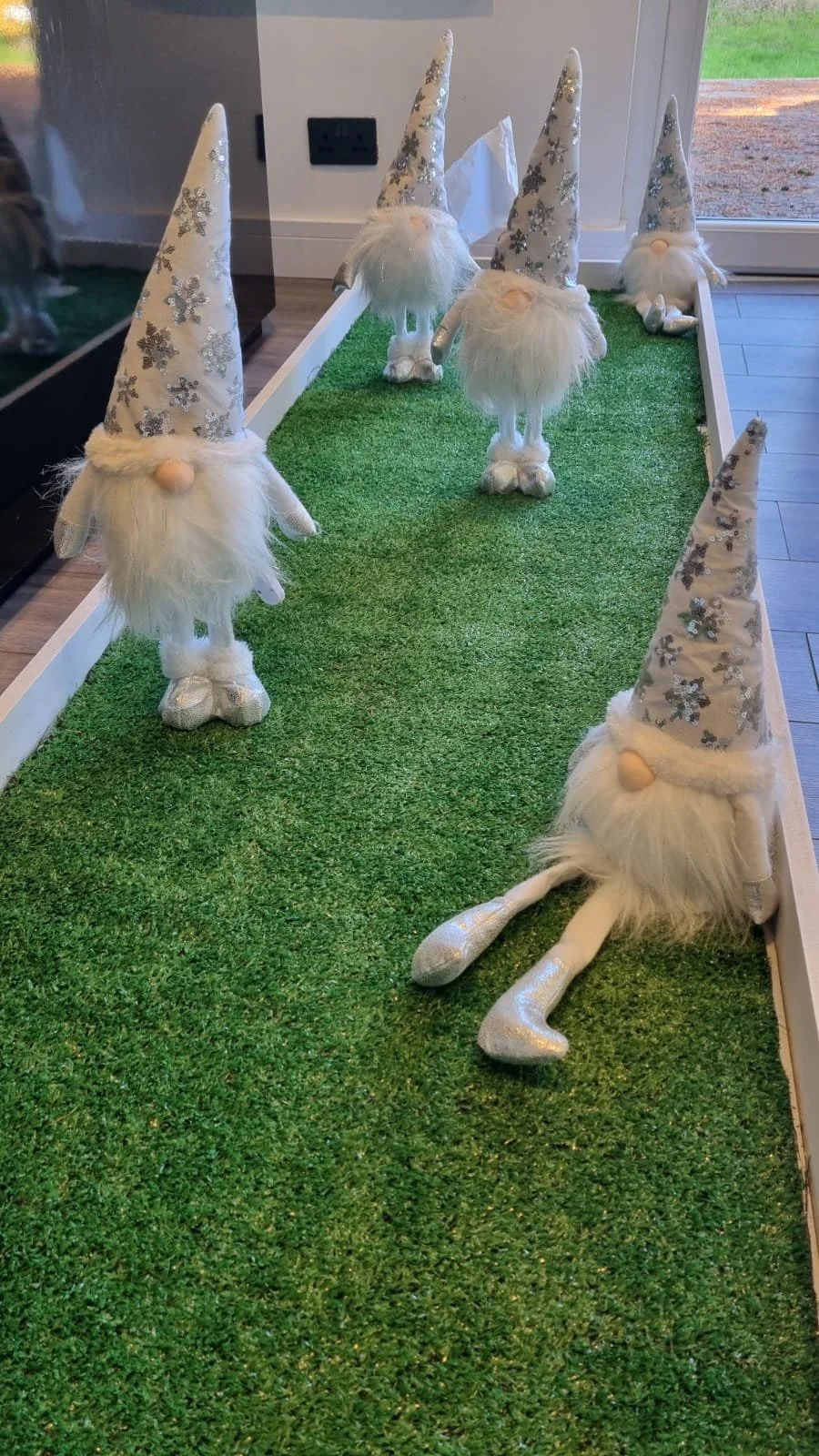 Decorative holiday display featuring five plush gnome figurines with long, pointed, snowflake-printed hats, white beards, and glittery shoes on a green grassy surface inside a house near a window.