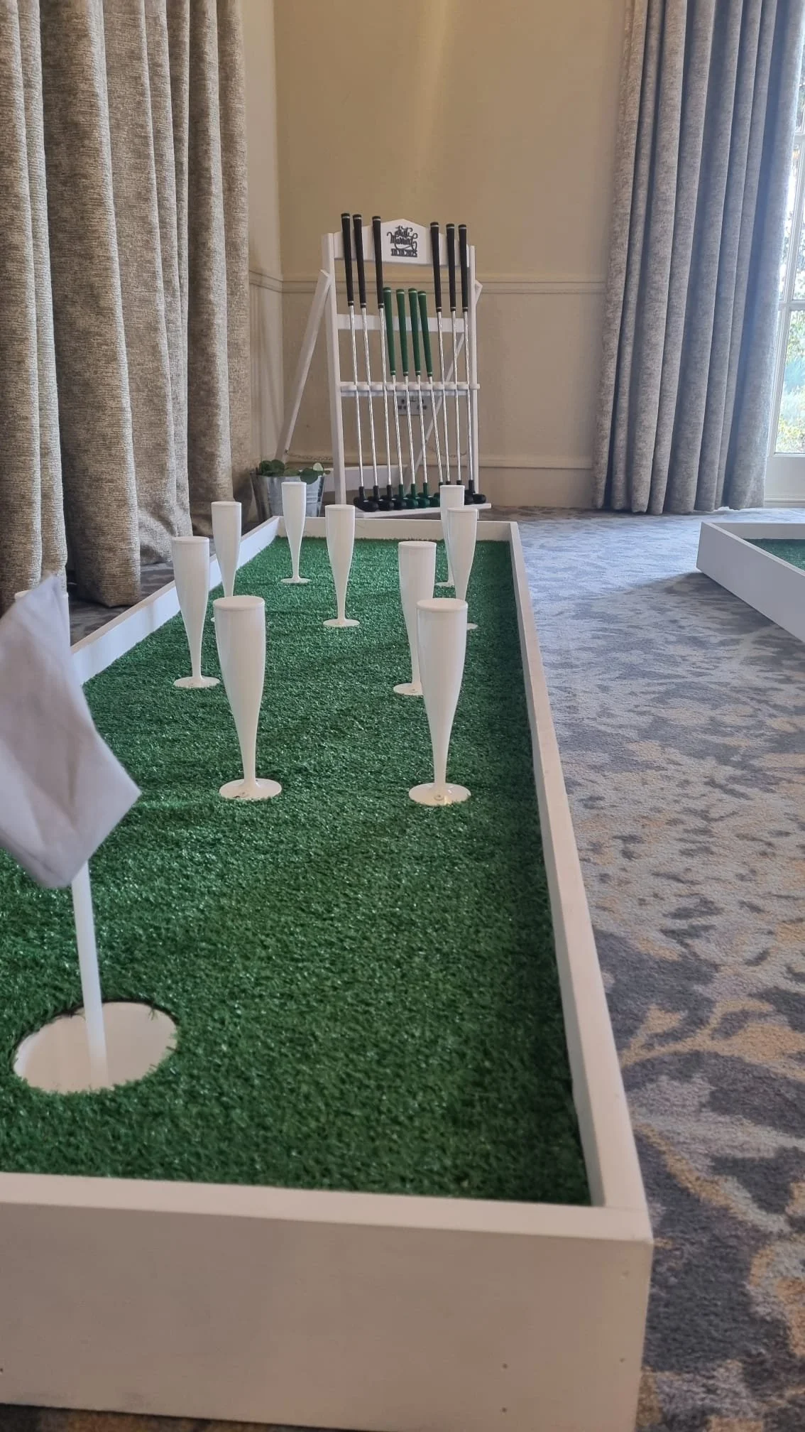 Miniature indoor golf game with a green turf, white golf holes, and small plastic golf clubs in the background.