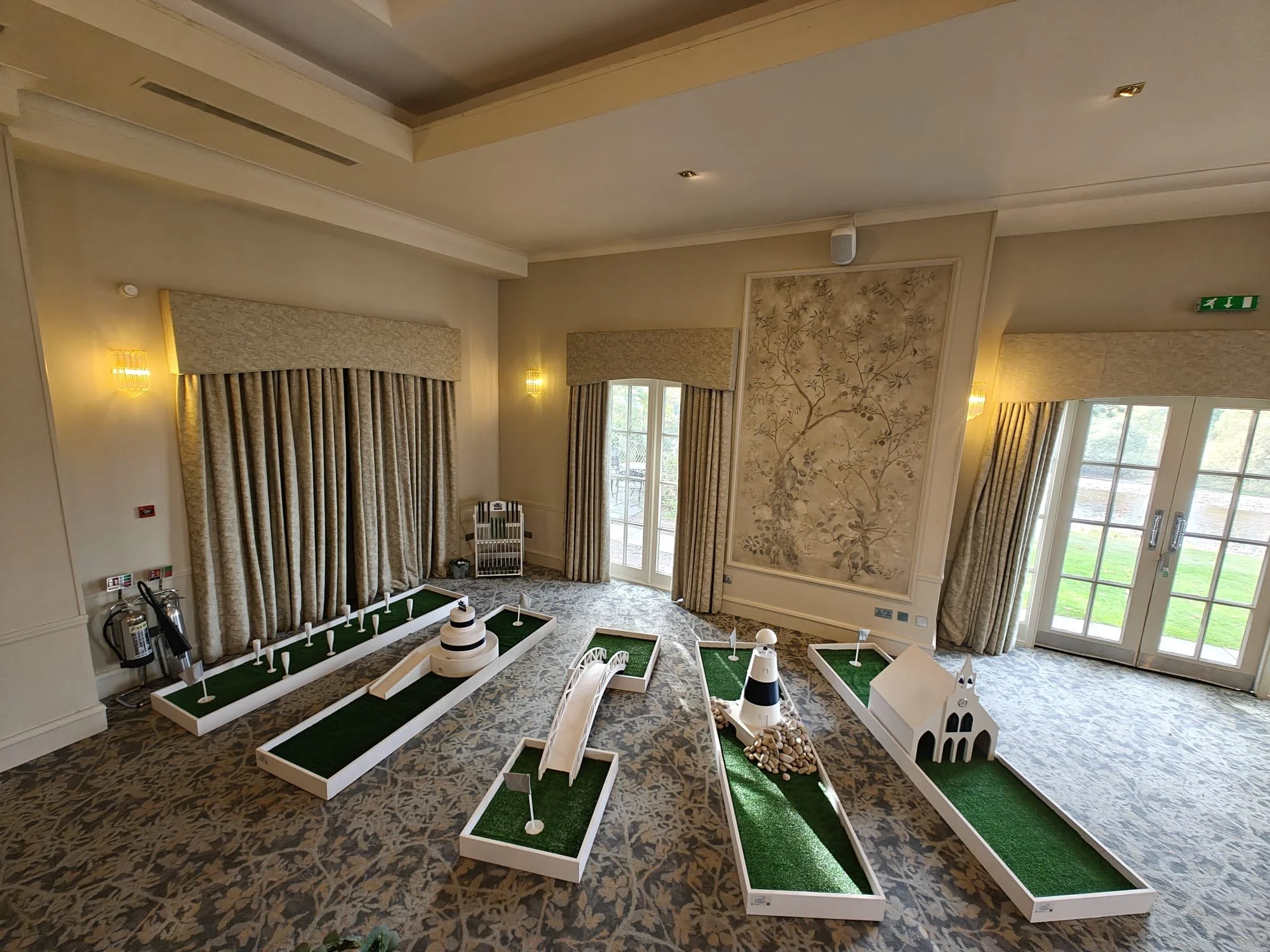 Miniature golf course setups in a luxurious indoor room with cream walls, a patterned carpet, large windows with curtains, and a decorative wall art. The setups include a lighthouse, a church, and various putting greens.