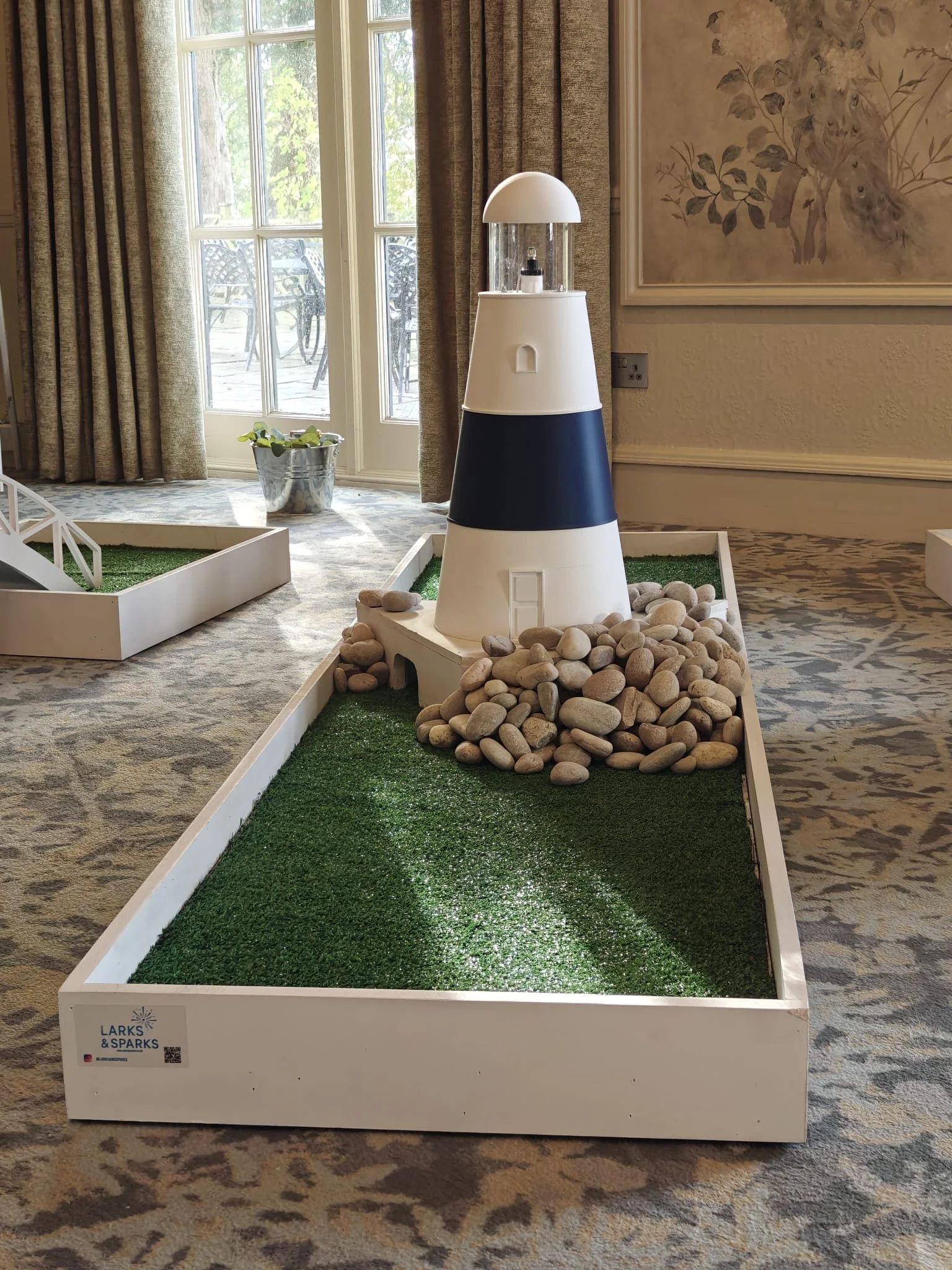 Miniature lighthouse model on a display base with rocks, artificial grass, and indoor decor.
