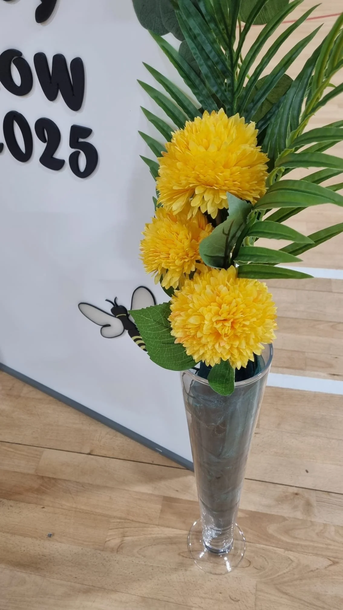 A tall glass vase containing yellow chrysanthemums, green leaves, and ferns, placed on a wooden surface. In the background, part of a whiteboard with black 3D letters and a bee illustration is visible.